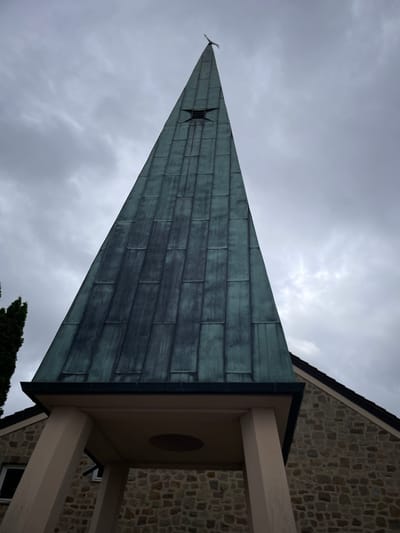 The image shows a low-angle view of the steeple of a building, likely a church, against a cloudy sky. The steeple is tall and pointed, covered in weathered green metal panels that form a series of vertical rectangles. Near the apex of the steeple, a dark, star-shaped opening is visible. At the very top, a thin, dark silhouette of a cross or bird is perched. The lower part of the steeple rests on a light-colored, open structure supported by pale pillars, with a stone building visible to the right. A portion of a tall, dark green evergreen tree is visible on the left side of the frame. The overall impression is one of a somber, possibly overcast day in Schorbach, Germany.