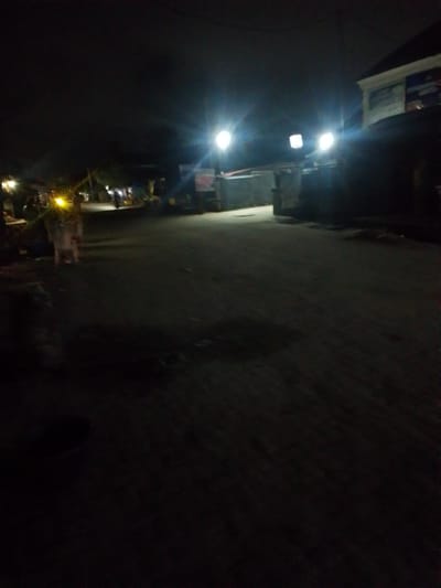 The image shows a poorly lit street at night in Awoyaya, Nigeria. Several streetlights illuminate the scene. Some people are present near the streetlights on the left side of the image. The road surface is visible, with shadows and uneven lighting. A building is visible on the right side.