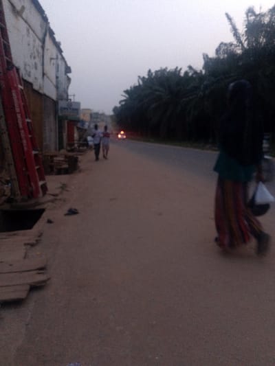 This image depicts a street scene in Abeokuta, Nigeria, at dusk or dawn under an overcast sky, characterized by low light. In the immediate foreground on the right, a person is walking away from the camera, wearing a dark head covering, a dark top, a long, vertically striped skirt in shades of red, brown, and green, and dark footwear. They carry a white bag in their left hand. The ground here is a light brown, unpaved surface transitioning to a darker, paved road in the midground.

On the left foreground, an open storm drain or trench is visible, partially covered by crude wooden planks. Red, vertical structures, possibly metal scaffolding or signage components, lean against a dilapidated building. This building, and others extending into the midground on the left, feature faded white and brown exteriors with shuttered windows or doors. A sign with indistinct text is visible on one building.

Further down the road on the left, two male figures are walking on the unpaved shoulder. Beyond them, blurred red and orange lights indicate distant vehicle headlights or taillights on the road. The road itself is a moderately wide, two-lane asphalt or paved surface. Along the right side of the road, a dense line of dark green palm trees forms a natural barrier, extending into the background. The sky is a uniform, hazy light gray.