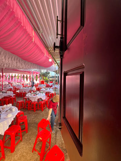 The image depicts an outdoor or semi-outdoor event space in Ko H'Neh, Vietnam, characterized by a festive setup for a large gathering. The scene is viewed from an angle partially obstructed by a dark, possibly mahogany, wooden door frame on the right, featuring a brass handle and a black latch mechanism.
The primary area is covered by a canopy structure with a pleated white ceiling, beneath which extensive pink fabric draping is suspended, adorned with small lights. Numerous round tables, each covered with a white tablecloth, are spread across the space. Many tables are visibly set with white ceramic dishes, bowls, and cutlery, suggesting preparation for a meal. Each table is surrounded by multiple bright red, backless plastic stools.
In the lower-right mid-ground, a person wearing a red short-sleeved shirt, blue jeans, and a light-colored bucket hat is bending over, interacting with one of the red stools, appearing to arrange or move it. Another individual in a light-colored shirt is standing near a table in the deeper background, toward the center-left. The ground beneath the tables and stools is an uneven, light-brown surface, possibly dirt or compacted earth. Green foliage and trees are visible beyond the event structure in the far background under natural daylight conditions.