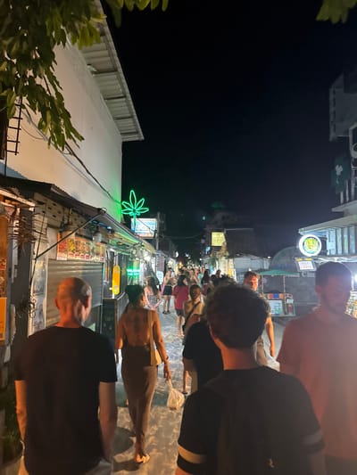 The image depicts a crowded nighttime street scene in Ban Ko Li Pe, Thailand. The narrow pedestrian thoroughfare is illuminated by artificial lights emanating from overhead fixtures and numerous commercial establishments lining both sides of the street. The sky above is dark.
In the immediate foreground, facing away from the camera, are four individuals. On the far left, a bald male in a black t-shirt walks with his back to the viewer. To his right, a female with dark hair, wearing a backless brown or tan ankle-length dress, holds a white plastic bag in her left hand and carries a light-colored shoulder bag on her right side. Directly in front of the camera, a person with dark, curly hair wears a black t-shirt with light horizontal stripes on the sleeves and a dark backpack. On the far right, partially visible, a male in a light peach or orange t-shirt walks.
Further down the street, dozens of other individuals, predominantly adults, are visible, walking in both directions, indicating a busy environment. The buildings lining the street house various shops and stalls. A prominent green neon sign shaped like a cannabis leaf is visible above a storefront on the left side of the street, approximately mid-frame. Other illuminated signs, including one with a Thai flag symbol and a round sign with green and yellow elements, are visible further down the street on the right. Skateboards are displayed vertically near a storefront on the left. The general environment suggests a bustling commercial or entertainment district.