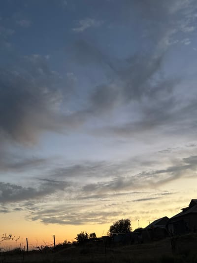 The image depicts a dusky sky over a rural settlement in Жібек жолы ауылдық округі, Kazakhstan. Silhouetted against the warm hues of the sunset, a cluster of modest structures, likely houses and outbuildings, are visible on a slightly elevated terrain. The foreground features a rustic fence made of thin posts and wire, intertwined with dry vegetation and thorny branches. The sky is a canvas of soft blues and grays, with wispy clouds stretching across, illuminated by the fading sunlight which casts a golden glow on the horizon. A single, faint star is visible in the upper right portion of the sky. The overall atmosphere is one of quiet twilight in a rural landscape.