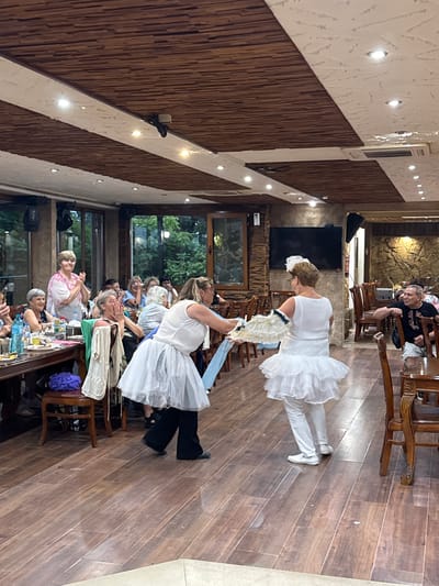 The image depicts an indoor scene, likely a restaurant or event hall, with a wooden ceiling and floor. The setting appears to be Sredets, Bulgaria, as indicated by contextual information.
In the foreground, two individuals are engaged in a performance or dance. One person, wearing a white layered skirt and white leggings, is positioned with their back to the camera, holding a fan. Another person, also in a white layered skirt and black pants, is facing the first individual, holding a blue sash or fabric. Their attire suggests a coordinated performance.
Surrounding them are several tables with diners, many of whom are looking towards the performers. There are bottles of drinks and plates on the tables. The diners are a mix of ages and genders, some appearing to be clapping or reacting to the performance.
In the background, there are more tables and chairs, along with windows that offer a view of greenery outside. The walls are decorated with stone and wooden elements, and a television screen is visible on one wall. Lighting fixtures are integrated into the ceiling. The overall atmosphere suggests a social gathering with entertainment.