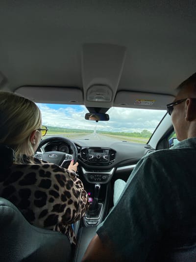 The image shows a view from inside a car, looking forward through the windshield. A woman with blonde hair and yellow-tinted glasses is driving, wearing a leopard-print top. Her hands are on the steering wheel. To the right and slightly behind her, a person wearing sunglasses and a dark t-shirt is seated. The car's dashboard and a gear shift are visible. The car is on a highway, with a scenic view of a rural landscape and a cloudy sky outside. There is no specific indication of the location being in the city of Niva, Russia, based on the visual information provided.