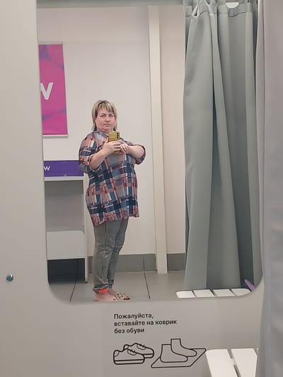 A medium-shot mirror selfie shows a woman standing in a fitting room. She is wearing a plaid shirt and striped pants. She is holding up a yellow smartphone to take the picture. The fitting room has grey curtains on the right side and a white wall on the left. In the background, a pink banner with the letter "V" on it is visible. On the bottom right of the image, there is text in Russian which translates to "Please, stand on the rug without shoes" and an illustration of shoes. The image appears to be taken in a retail store.
