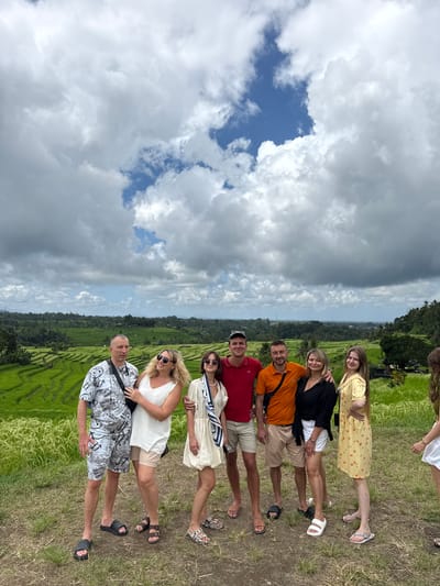 A group of seven people stands in a line on a grassy area, seemingly posing for a picture. Behind them is a landscape with rice terraces and some trees. The sky is visible above, filled with clouds. The location appears to be in Penebel, Indonesia. The individuals vary in age and wear casual clothing suitable for warm weather. Some are dressed in shorts and t-shirts, while others wear dresses or shirts with shorts. They seem to be tourists enjoying the scenery.