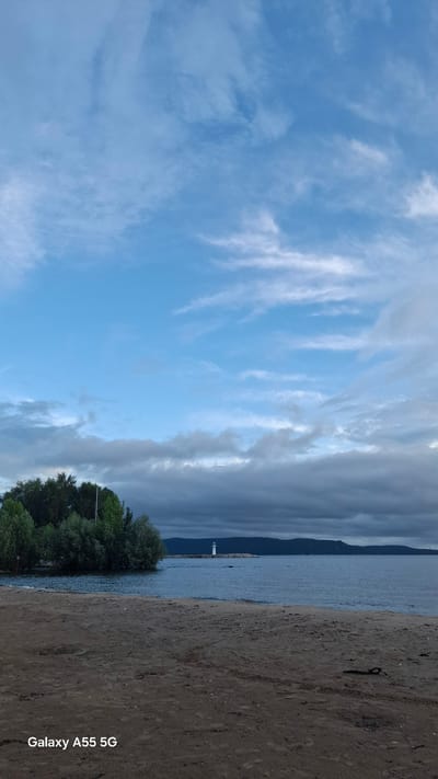 This image depicts a scenic view of a beach and a body of water, likely a lake or sea, under a sky filled with wispy clouds. The foreground is dominated by a sandy beach, with a dark, textured surface. To the left of the beach is the calm water, and in the distance, a faint outline of mountains or hills can be seen. A solitary white lighthouse stands on a pier or landmass jutting into the water. A cluster of green trees is visible to the left, near the water's edge, with some foliage extending towards the lighthouse. The sky is a vibrant blue with streaks of white clouds, suggesting either early morning or late afternoon. The overall mood is peaceful and serene. The image also features a watermark in the lower right corner that reads "Galaxy A55 5G".
