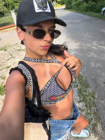 A young woman is pictured outdoors, wearing a black baseball cap with a black panther patch, round sunglasses, a patterned bikini top, and ripped denim shorts. She is holding the strap of her bikini top with her right hand. A straw bag with black fringe is visible next to her. She is wearing light blue slide sandals on her feet, one of which is visible. The scene appears to be a sunny day, with shadows cast on the asphalt path she is sitting on. Green foliage and gravel are visible in the background. There is no visible text. The location is Chavei, Bulgaria.
