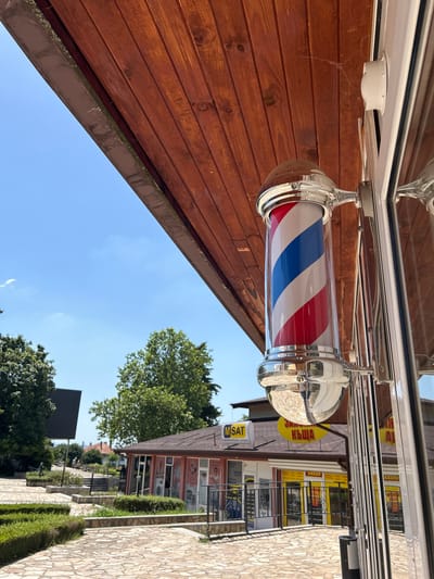 This media file displays an outdoor scene featuring a traditional barber pole with red, white, and blue stripes, suspended from a white awning. The barber pole is a cylindrical glass tube with chrome accents. The background reveals a building with shops, some with yellow awnings and signage. One sign is partially visible with Cyrillic text. A ladder is leaning against one of the buildings. To the right, a stone-paved walkway runs alongside manicured green hedges. Trees and a clear blue sky with a few wispy clouds are visible above the buildings and walkway. The image appears to be taken on a sunny day. Visible text includes "MSAT". The orientation of the image is rotated.