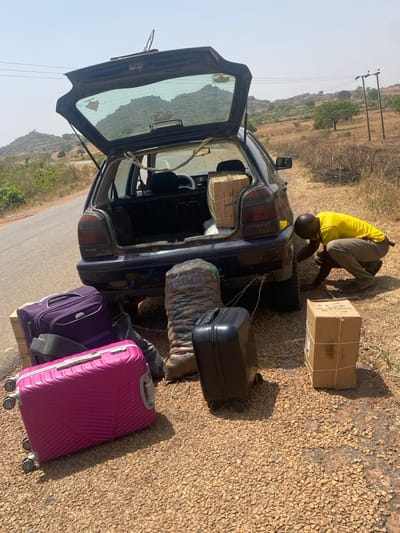 This image, taken in Zankan, Nigeria, depicts a dark-colored hatchback car parked on a gravel shoulder adjacent to an asphalt road under a clear, bright sky. The car's trunk is open, revealing a large, rope-secured cardboard box and other contents. A single adult male, wearing a yellow short-sleeved shirt and light-colored trousers, is in a crouched position near the car's rear-right wheel, appearing to inspect or attend to something beneath the vehicle. Scattered on the gravel around the car are several items of luggage and cargo: a large purple wheeled suitcase, a large pink wheeled suitcase, a smaller black wheeled suitcase, a tall burlap sack labeled "PORKOS," and a rope-bound cardboard box. The background features sparsely vegetated hills and power lines with poles.