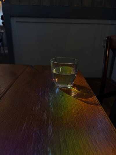 A clear glass of water sits on a wooden table, casting a rainbow of light onto the surface. The table appears to be in a dimly lit room, possibly a bar or restaurant, given the presence of a wooden chair in the background and the dark, textured wall behind it. The lighting creates a dramatic effect, highlighting the water in the glass and the grain of the wood. The scene is still and quiet, suggesting a moment of contemplation or a pause in activity. The location is specified as Ipswich, United Kingdom.