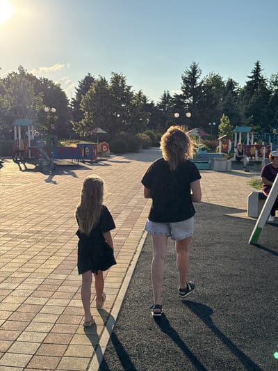 The image shows two people standing on a paved area next to a playground. The person in the foreground, seen from the back, has long, blonde hair and is wearing a black dress and light-colored sandals. The second person, further back and also seen from the back, has curly, brown hair and is wearing a black t-shirt and denim shorts with sandals. The sun is setting, casting long shadows across the ground, suggesting it is late afternoon or early evening. The sky is clear with some wispy clouds. The setting appears to be a public park or recreational area, with playground equipment visible in the background. The overall atmosphere seems relaxed and casual. There is no visible text in the image.