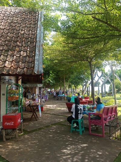 A bustling outdoor space features vendors under a tiled roof. People are seated at colorful tables, enjoying the shade beneath tall, leafy trees. A woman in a black and white jersey sits on a green stool, while another woman sits on a pink bench. A red Coca-Cola cooler is visible near one of the vendor stands. Decorative, abstract structures are seen in the distance.