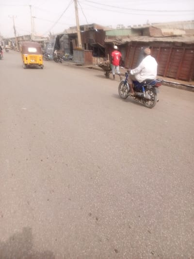 The image captures a daytime street scene, likely in an urban or semi-urban environment, characterized by a hazy or dusty atmosphere. A wide, light-colored asphalt road occupies the foreground and midground of the image.
On the right side of the road in the midground, a man wearing a white shirt or tunic and light-colored trousers is riding a blue motorcycle, moving away from the camera. To his left, another man, dressed in a red t-shirt with the number "1" prominently displayed on the back and a white cap, is pushing a wooden wheelbarrow. The wheelbarrow appears to contain various items. Behind these individuals, a row of informal structures, possibly small shops or stalls, line the roadside. These buildings are constructed predominantly from corrugated metal sheets and wood, with some featuring closed wooden doors or shutters.
On the left side of the road, a yellow three-wheeled auto-rickshaw, seen from its rear, is also moving away from the camera. Further down this side of the street, other vehicles, possibly motorcycles or more auto-rickshaws, are visible, either parked or in transit. Similar makeshift structures also line this side of the road.
The background is somewhat blurred and indistinct due to the hazy conditions, revealing more buildings and numerous utility poles with a dense network of overhead electrical or communication wires, extending into the distance. The sky is bright but appears overcast or dusty, consistent with the overall hazy quality of the image. The scene depicts typical daily activity on a community street.