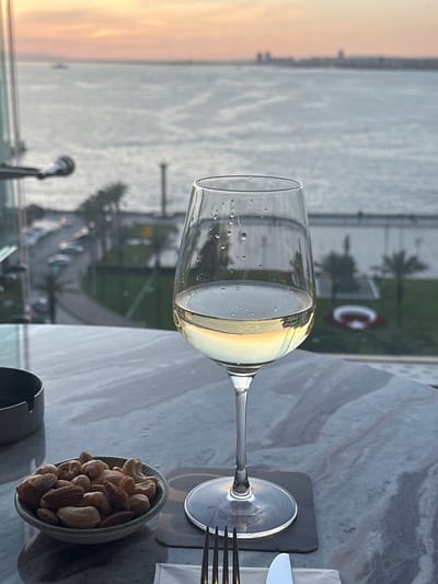 A table is arranged in a high-altitude location, seemingly a restaurant or a similar establishment, offering a panoramic view of what appears to be the city of İzmir, Turkey, alongside a body of water. The focus is a wine glass filled with a light-colored liquid, likely white wine, adorned with condensation droplets. The reflection in the wine glass shows a sunset over a body of water and buildings. The outside scene includes a view of water, buildings in the distance, and a visible Turkish flag created in what appears to be landscaping. On the table, there's a small bowl filled with mixed nuts, a napkin, and cutlery. The tabletop has a gray marble pattern, implying an upscale venue.