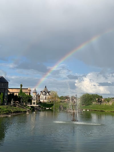 The image depicts a scenic view of a park or amusement area located in Malakhovetsky Selsoviet, Belarus. A prominent rainbow arches across a cloudy sky, with a double rainbow faintly visible above the main arc. In the foreground, a calm body of water features a fountain actively spraying water upwards. Along the bank of the water, several white swans are visible. The background showcases a collection of charming, whimsical buildings with European-inspired architecture, reminiscent of a fairytale village. Green grassy areas and trees surround the structures and the water body, contributing to the picturesque atmosphere. The overall impression is one of a serene and visually appealing landscape.