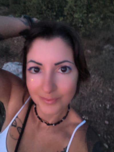A frontal, waist-up selfie depicts an adult female outdoors in Shaalvim, Israel, under soft, low-light conditions, likely dusk. The subject has dark brown hair styled short, dark brown eyes with prominent blue eyeliner on both upper and lower lash lines, and a small silver nostril piercing on her right nostril. Her skin appears warm-toned, illuminated by a gentle, reddish glow, with a noticeable glitter or highlight on her left cheekbone near the eye. She wears a white spaghetti-strap top, partially revealing dark, abstract tattoos on both shoulders/upper arms. A black beaded choker necklace with silver metallic accents is around her neck. Her left arm is raised, with her hand positioned near her head/hair, while her right arm is mostly out of frame. She gazes directly into the camera with a subtle, closed-mouth smile. The background is out-of-focus, showing dark green foliage, dry earth, and scattered rocks, indicating an undeveloped outdoor setting.