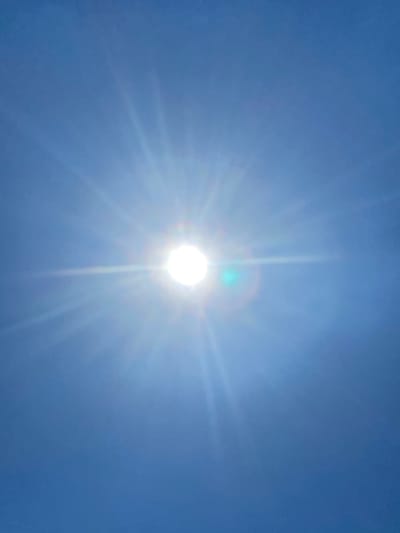 The image is a close-up, vertically oriented shot of the sun in a clear blue sky. The sun is the dominant feature, positioned slightly right of center, and appears as a bright, white disc. Sun flares radiate outwards from the sun, creating a starburst effect that fills much of the frame. The sky is a consistent, deep blue, with no clouds visible. There are some faint lens flares, including a noticeable green and blue circular flare to the right of the sun. The overall impression is of a very bright, sunny day. The location provided, Matiashivka, Ukraine, is an external environmental context for this image, but the image itself shows no discernible landmarks or features specific to any city or location.