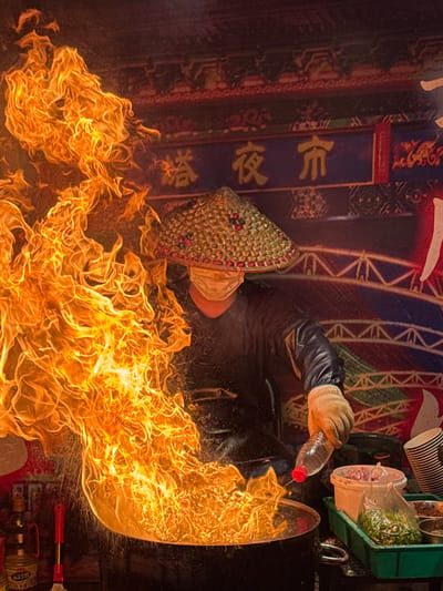 The image depicts a street food vendor in Guta, China, preparing a dish over a large flame. The vendor, wearing a conical straw hat and a face mask, is actively cooking in a wok, with intense flames erupting from it. A plastic bottle of liquid is held by their gloved hand, suggesting they are adding it to the wok. Several containers with chopped ingredients, possibly vegetables and spices, are visible nearby, along with a stack of paper cups. The background features a colorful banner with Chinese characters, likely advertising the night market. The overall scene captures the dynamic and vibrant atmosphere of a bustling Chinese night market.
