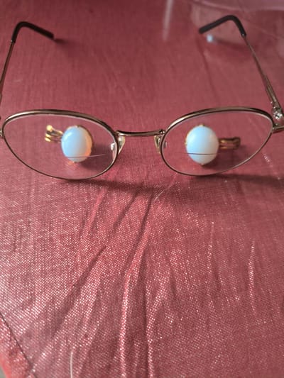 The image shows a pair of eyeglasses with round lenses resting on a textured, red fabric surface. Each lens has a small, white, oval-shaped object attached to the inside, appearing to be decorative charms or perhaps weights. The eyeglasses are silver-toned metal with black tips on the temples. The fabric surface exhibits a crinkled texture and catches the light, suggesting it might be silk or a similar material with a subtle sheen. The background is out of focus, but the overall impression is of an indoor setting. There is no indication of the location being in Sokury, Russia, as this information is not present in the visual data.