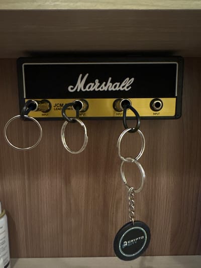 The image shows a wall-mounted key holder designed to resemble a Marshall amplifier head. The key holder is black with a gold faceplate, and features the "Marshall" logo in white script. There are three input jacks, labeled "INPUT" and "JCM LEAD SERIES".
Three keychains are hanging from the input jacks. The top keychain has a black loop that connects to two silver key rings, which are linked together in an infinity symbol shape. The middle keychain has a black loop connected to a single silver key ring. The bottom keychain has a black loop connected to a larger silver key ring. Attached to the top keychain is a round, black fob with "CRYPTO CITY" printed in white text within a blue and white circular border.
The key holder is mounted on a wooden surface, likely a wall or shelf, with a horizontal wood grain pattern. The lighting suggests an indoor environment. The image is cropped to focus on the key holder and keychains. There is no indication of the time of day, weather, or any emotions. The location context is provided as Orsk, Russia, but there are no visible cues in the image to confirm this.