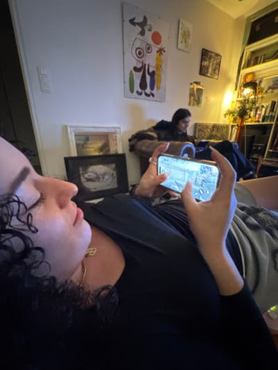 The scene depicts an indoor living area in Vincennes, France, occupied by two individuals. In the foreground, a person with dark, curly hair is positioned on their back, viewed from the mid-chest upward, wearing a dark long-sleeved top and a gold chain with a small, light-colored pendant. Their eyes are closed or cast downwards. Both hands hold a smartphone horizontally, displaying an active mobile video game with a landscape and user interface elements. The person's lower body is partially covered by a patterned grey and white blanket.
In the background, a second person with dark hair is seated on a brown armchair or sofa, obscured from the waist down, appearing to interact with a personal device in their lap. They are wearing dark clothing.
The room features light-colored walls adorned with several artworks: a large, vertical abstract poster with colorful shapes (red circle, brown figures, elongated yellow/green forms), a smaller framed landscape painting with a white frame, and a dark-framed monochrome image below it. A white light switch is visible on the wall. To the right, a tall white bookshelf filled with books and various items, including a small plant in a vase and a light fixture, is present. The overall lighting is dim, supplemented by light emitted from the smartphone screen and the bookshelf light. No direct interaction between the two individuals is evident.
