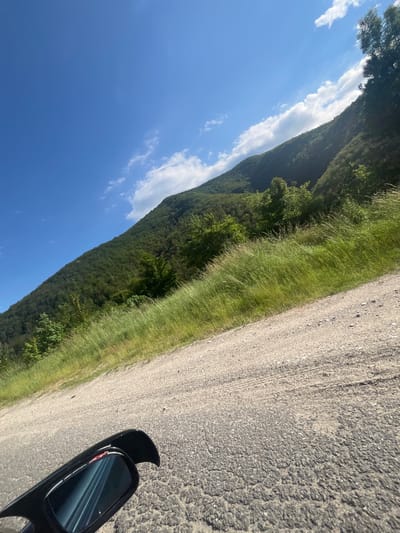 The image depicts a tilted view of a mountainous landscape on a sunny day, likely in or near the city of Taran, Bulgaria. A dark side-view mirror of a vehicle is visible in the foreground on the lower left, reflecting a blue sky and what appears to be a road. The road surface is a mix of asphalt and gravel, with visible tire tracks. To the right of the road, tall green grass grows, and beyond that, a densely forested mountainside rises steeply. The sky is a vibrant blue with scattered white clouds. The overall impression is one of a scenic drive through a rural or mountainous region.
