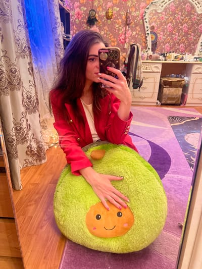 A young woman with long brown hair is taking a selfie in a room. She is wearing a red blazer over a white top. In front of her is a large, plush avocado toy. The room has floral wallpaper and is lit by a blue light from the left and fairy lights in the background. To the right, a vanity dresser is visible with various items on top. A mirror reflects the scene, showing the woman and the room. The floor is wooden, covered in part by a purple rug with abstract patterns. The image is a personal snapshot taken in a domestic setting, likely a bedroom or dressing area, and there are no specific indicators of the city of Krasnoe-na-Volge, Russia.
