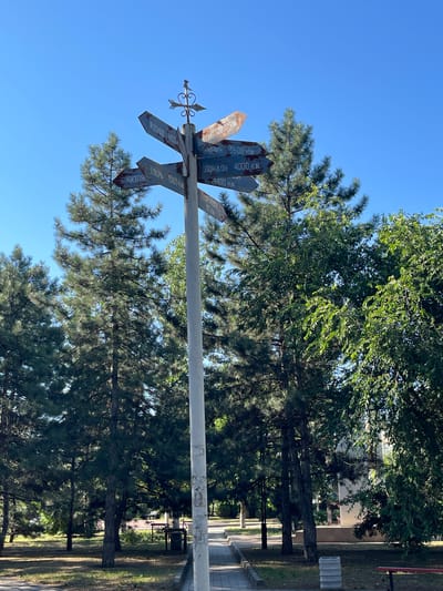 The image shows a directional signpost in the city of Pokoinoe, Russia. The signpost, made of white metal, has multiple arms pointing in different directions, each with a rusted sign indicating city names and distances in kilometers. For example, one sign reads "МОСКВА 1450 KM" (Moscow 1450 KM), another "Лондон 4000 KM" (London 4000 KM), and "ТВЕРЬ 1565 KM" (Tver 1565 KM). The signpost is topped with a decorative metal element resembling an arrow and a heart. It is situated in a park-like setting with several tall pine trees and deciduous trees surrounding it. A paved walkway runs in front of the signpost. The sky is clear and blue. In the background, parts of buildings and other park benches are visible. There are no people or animals in the image.