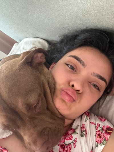 This close-up media file features a young woman and a brown dog. The woman has dark hair, dark eyebrows, and is making a kissing face, puckering her lips. She is wearing a white top with a floral pattern in shades of pink and red. The dog appears to be a pitbull breed, and it is snuggled up close to the woman, with its head resting on her neck or shoulder. The dog's eyes are closed, suggesting it is relaxed or sleeping.
The scene appears to be indoors, possibly in a bed or on a couch, given the presence of blankets or soft surfaces. The lighting suggests it could be daytime, but no specific time can be determined. The overall emotion conveyed is one of affection and comfort between the woman and her pet. The image is taken from a high angle, looking down on the subjects. There is no visible text in the image. The location context provided is Malevo, Bulgaria.