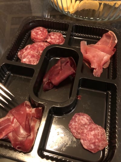 The image shows a black plastic charcuterie tray containing different types of cured meats. There is a pile of thin, pink prosciutto in the top left section, and a couple of slices of speckled salami in the top right section. The central section contains a stack of dark red bresaola, and the bottom left section holds several small rounds of chorizo. The bottom right section features more prosciutto. A fork is partially visible in the bottom right corner. The tray is placed on a reflective surface, likely a glass table, with a blurred glass object visible in the upper left corner. No people or specific location details pertaining to Kehrsatz, Switzerland are discernable from this image.