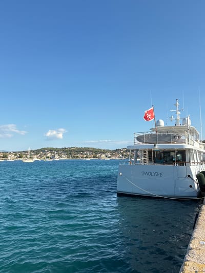 In Limenas Markopoulou, Greece, a modern white yacht named "PAOLYRE VALLETTA" is docked alongside a stone pier. The yacht is viewed from its stern, with the name clearly visible on the hull. A red flag with a white cross is flown from the mast. The water in the foreground is a deep blue, showing ripples from the gentle movement of the sea. In the background, a coastline dotted with green trees and white buildings stretches under a clear blue sky with a few scattered clouds. Several other sailboats are anchored in the bay. The overall scene depicts a serene coastal environment.