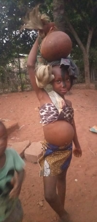 A young girl in traditional attire is carrying a large gourd on her head. She is wearing a leopard print top and a patterned skirt. Her face is partially painted black. A fair-haired wig is draped over one arm. A blurred figure of a young child is partially visible in the lower left corner. The setting appears to be a rural outdoor area with red dirt and scattered vegetation. The image is taken in dim lighting.