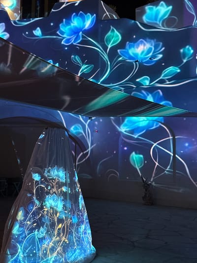 The image shows an art installation, likely a projection mapping display, with blue floral designs and white line art covering multiple surfaces, including what appears to be fabric draped over a structure. The scene is interior and displays a modern or contemporary aesthetic. There is no explicit evidence to confirm the location is specifically in Ghadeer Al Tayr, United Arab Emirates. There are no people directly visible in the image. The environment consists of structural elements with the projection displayed on them.