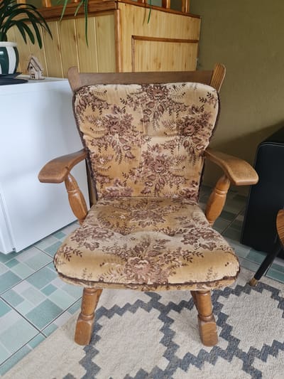 An antique wooden armchair with a floral upholstered seat and back is positioned on a patterned rug. The chair has carved wooden arms and legs. Behind the chair is a light-colored cabinet made of wood planks, and to the left is a white appliance, possibly a refrigerator. The floor is tiled with a geometric pattern of light blue and white squares. In the upper left corner, a plant in a green and white pot is visible, along with a small decorative house. The wall behind the chair is painted a muted olive green. The scene appears to be indoors, in what might be a residential setting in Molėtai, Lithuania. There are no people or animals present.