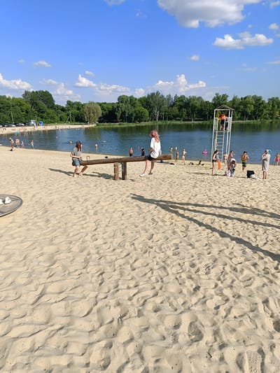 In Gubkin, Russia, a bright, sunny day at a sandy beach near a lake is captured. Several individuals are present on the beach and in the water. In the foreground, a seesaw made of wood is set up on the sand. Two young people are seated on the seesaw. One, a person with pink-tinted hair wearing a white t-shirt and black shorts, sits on one end, while another person with dark hair wearing a grey t-shirt and blue shorts sits on the other. To the left, a circular metal structure, possibly a part of a playground, is visible on the sand. In the background, a lake stretches out, with trees lining its far bank. Scattered across the beach are various beachgoers, some wading in the water, others relaxing on the sand, and a few playing near the shore. A lifeguard tower stands prominently near the water's edge. The sky is a clear blue with scattered white clouds.