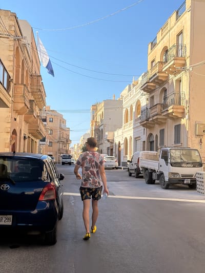 A man walks away from the camera down a narrow, sun-drenched street in Xagħra, Malta, on a clear day. The man is wearing a floral print t-shirt and patterned shorts, with yellow shoes. He is carrying a clear plastic bottle in his right hand. To his left, a dark blue hatchback car is parked. The street is lined with traditional Maltese buildings featuring stone facades, arched windows, and decorative balconies with wrought-iron railings. The buildings are predominantly in a pale yellow or cream color. Overhead, electrical wires are strung across the street. In the distance, other vehicles are visible, including a white van and a white ISUZU flatbed truck with a license plate ending in "446". A flag with white and blue sections is flying from a building on the left side of the street. The road surface is asphalt with a white line marking the center.