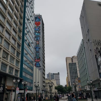 The image shows a street scene in Curitiba, Brazil, with a focus on the architecture and street life. The sky is overcast. Buildings of varying heights dominate the view. One building is distinguished by a colorful graffiti mural reading "EU AMO CURITIBA" (I LOVE CURITIBA). The building has a grid-like pattern of windows. People are walking along the sidewalk, some standing and interacting. Street lamps are visible, lining the edge of the sidewalk. Signs and store fronts are also visible at ground level.