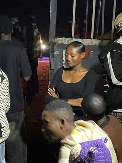 In Wumat, Nigeria, a group of individuals are gathered near an opening in a fence. A young woman with her hair pulled back and a few red strands visible is looking down and to her right, her arms crossed. She is wearing a black, long-sleeved shirt and a small necklace. To her left, a person wearing a black shirt and jeans is partially visible from the back. To the right of the woman, another person wearing a black jacket with white stripes is standing. In the foreground, two individuals are partially visible. One is wearing a light-colored jacket over a brown garment and a purple crocheted hat with white and yellow accents. The other person is seen from the side and is wearing a brown garment. The background appears to be a dirt path with some lights visible in the distance. The scene has a dim, nighttime ambiance, possibly indicating an outdoor event or gathering.