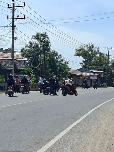 The image, rotated 90 degrees clockwise, depicts a roadside scene in Langa, Indonesia, under a bright, clear sky. A line of approximately seven to eight motorcycles, most with riders in helmets, is stopped or parked along the right side of a paved road. Many individuals are seated on their bikes, while some stand beside them. The road features a solid white line delineating its edge.

To the right of the road, behind the parked motorcycles, are several modest buildings with corrugated metal roofs, interspersed among verdant trees. One visible structure has signage that clearly displays "POLO" and "Barber," suggesting a local business. Numerous utility poles carrying a dense array of power lines stretch across the left side of the frame and into the sky. The sharp shadows visible on the ground indicate it is midday or early afternoon. The scene captures a momentary halt or gathering of motorcyclists along a local thoroughfare.