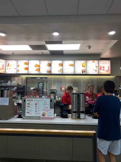 The image depicts the interior of a Chick-fil-A restaurant, likely in Hagerstown, Maryland, United States of America, based on the visible branding on an employee's shirt. The scene is a counter area where customers interact with staff.

In the foreground, a young male wearing a dark blue t-shirt and light-colored shorts stands facing away from the camera, looking towards the counter. Behind him, a counter separates the customer area from the food preparation space. On the counter, there are various items including a credit card reader, a menu board displaying "Kids" meals, entrees, sides, and drinks, and a "Original Sandwich $2.50" sign. There are also two tall, stainless steel beverage dispensers.

Behind the counter, three young female employees are visible. One, in a red shirt, is interacting with a customer or colleague. Another, also in a red shirt with "CHICK-FIL-A HAGERSTOWN EST. 2001" printed on it, is looking towards the camera and appears to be a cashier or server. A third employee, in a red shirt, is partially visible further back. The wall behind the counter is covered with a textured, light-colored tiled backsplash. Above the counter, a menu board displays various food and drink options, categorized by "Meals Include," "Original Meals," "Grilled Meals," "Salads," "Drinks," "Treats," and a special offering of "Pineapple Dragonfruit." The ceiling features recessed lighting and acoustic tiles. A security camera is visible mounted on the ceiling. The overall environment suggests a typical fast-food restaurant setting during operational hours.