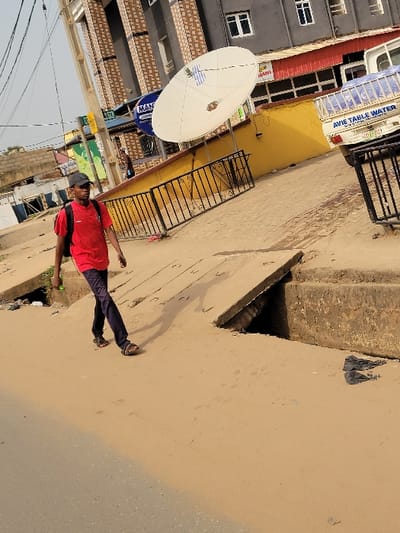 The image displays an urban street scene in the city of Puerto López, Colombia, under bright daylight conditions. In the foreground, a male individual, appearing to be an adult, walks from left to right across a sandy, light brown pathway. He wears a red short-sleeved t-shirt, dark trousers, and dark sandals, carrying a black backpack and a black baseball cap. His shadow is visible to his right on the ground.
The immediate ground level consists of packed sand and dirt, transitioning to a dark asphalt road surface on the far left. To the individual's right, several uneven concrete slabs form part of a broken or incomplete drainage system, revealing dark openings beneath them. Black plastic debris is present on the sandy surface near these concrete structures on the right side of the frame.
In the mid-ground, a black metal fence with decorative elements separates the pathway from a yellow-painted wall. A large white satellite dish with blue and white markings is mounted on this yellow wall. To the left of the dish, a round blue sign with "MAN Power Lounge" written in white text is visible. Further left, a concrete utility pole with overhead wires extends upwards.
Behind the fence and yellow wall, a multi-story building with a grey facade and brown textured vertical sections is visible, featuring multiple white-framed windows. A red awning or roofline is present over a lower section of a building behind the satellite dish. On the far right, a white pickup truck, marked with "AVIE TABLE WATER" on its side, is partially visible, parked beyond the yellow wall. A second, less distinct individual is partially visible behind the black fence and blue sign in the mid-background.