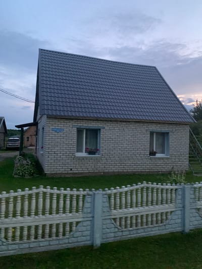 The image shows a small, light-colored brick house with a gray, sloped roof. The house has two windows on the visible side, each with a planter box. A street sign reading "СОВЕТСКАЯ 25" is mounted above the left window. A white concrete fence with decorative balusters runs across the foreground. Behind the house, a portion of a dark-colored vehicle is visible, along with a wooden structure and some vegetation. The scene appears to be in a rural or suburban setting, possibly a village, given the street sign and the overall architecture. The sky is overcast with some clouds, suggesting it might be late afternoon or early evening. The location is identified as Відзаўскі сельскі Савет, Belarus.