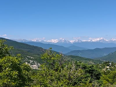 The image depicts a panoramic view of a mountainous landscape with a clear blue sky. In the foreground, lush green trees, some with bare branches, frame the scene. Beyond the trees, rolling hills covered in dense green forests recede into the distance. Several layers of mountains are visible, with the furthest peaks capped with snow. The time of day appears to be daytime, with bright sunlight illuminating the landscape. The weather seems to be clear and pleasant. The scene evokes a sense of natural beauty and tranquility. Hints of human settlement can be observed in the distance, with small houses nestled on the hillsides. The overall setting is a rural mountainous region, consistent with the provided location context of Ukeshi, Georgia. No people, objects, text, or specific activities are clearly identifiable.