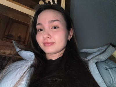A close-up indoor portrait, likely a selfie, depicts a young woman in Bayronovka, Russia. The subject, positioned center-right, has long dark hair and fair skin, looking directly at the camera with a neutral expression and a slight smile. She is wearing a dark, possibly black, inner garment beneath a light blue quilted or puffer-style jacket. A partial white logo, resembling "ornaments," is visible on the jacket's right side, near the shoulder. Her visible right ear features a small hoop earring and a separate stud earring. The background on the left displays dark wooden paneling, including a lighter horizontal wooden beam at the top and a recessed wooden section below. The background on the right is a plain, dark grey wall. The scene is dimly lit, with the subject's face being the most illuminated area.