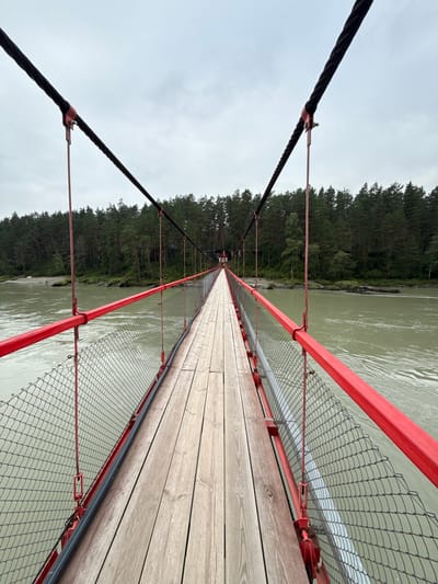 An eye-level, full shot shows a perspective of a suspension bridge leading across a wide, murky river. The bridge has wooden planking for its walkway and is lined with bright red railings and chain-link fencing. Thick, black cables run along the top of the bridge, supported by red poles. The river flows with a greenish-brown hue, and on the opposite bank, a dense forest of dark green coniferous trees stretches across the horizon. The sky above is overcast and a pale gray. The scene is likely located in or near the village of Barangol, Russia, given its natural setting with a river and forest. There are no people visible on the bridge or in the immediate vicinity.