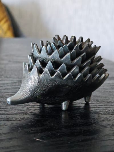 A close-up, eye-level, medium shot shows a metallic hedgehog figurine placed on a dark, horizontally grained wooden surface. The figurine is dark gray with subtle green accents near the eyes, and its spines are layered and pointed, resembling saw teeth. The hedgehog is facing left, with its snout extended, and its three visible feet are small and rounded. The background is softly blurred, with a lighter area on the left and a light gray wall on the right. The lighting is bright and even, casting a soft shadow behind the figurine. The location in the city of Заклинье, Russia is not identifiable from the image.