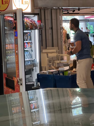 This image depicts an indoor scene in San Cristóbal de La Laguna, Spain.
In the left foreground, a white and orange vending machine is present, with its right service door open. The machine's glass front displays rows of bottled beverages (e.g., Fanta, water). The open section reveals internal shelving stocked with bags of crisps/chips on the top shelf and various canned/bottled drinks (e.g., K-Tonic, water) on shelves below. Above the vending machine, an illuminated sign displays "1,50" in red and orange text.
To the right, a male individual is visible from the mid-torso to the lower legs, facing left. He has short, dark hair and a beard/stubble. He wears a blue short-sleeved t-shirt, light beige trousers, and a dark-colored smartwatch on his left wrist. His hands are slightly cupped, positioned as if holding or preparing to handle an item. On the floor in front of him are two dark blue plastic crates containing various cardboard boxes and packing materials. Behind these crates, several white chairs are stacked in a disorganized manner, alongside a light-colored table.
In the background, an accordion-style security grate is partially visible, behind which an open doorway leads to an exterior or brightly lit internal area. A female individual, viewed from behind, wearing light-colored patterned trousers and a dark backpack, stands near this doorway. To her right, a second male individual wearing a white shirt is partially visible in the background.
The lower third of the image consists of a clear, inverted reflection of the vending machine, the blue crates, and the lower half of the primary male subject, indicating a highly reflective floor or surface in the immediate foreground.