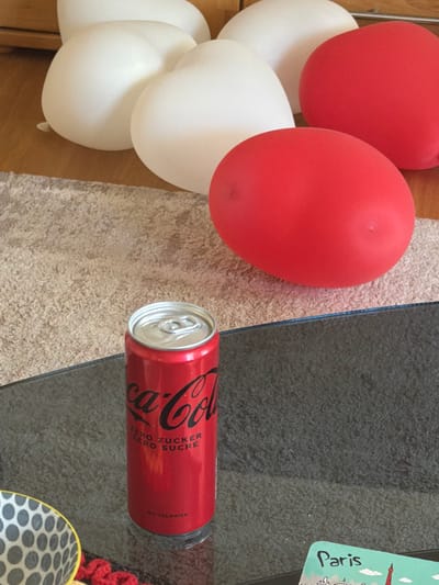 A can of Coca-Cola Zero Sugar, with German and French writing, sits on a glass-topped table in Basel, Switzerland. Next to the table, on a beige carpet, is a bunch of white and red balloons. Part of a wooden floor is visible in the lower-left corner. A postcard with an image of Paris is visible on the top-right.