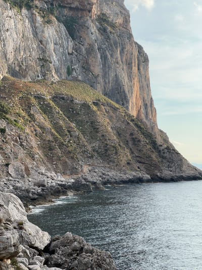 This media file captures a dramatic natural scene of a steep, rocky mountainside meeting a large body of water. The mountain is rugged and textured, with layers of rock exposed and varied shades of gray and tan. Patches of dry grass and sparse vegetation cling to the slopes, particularly on the middle section of the mountain. The water on the right side of the frame is vast and appears to be choppy, with a textured surface reflecting the light, suggesting it is a sea or a large lake.
The perspective is from a high vantage point, looking down and across the landscape. The time of day appears to be daytime, with a bright, slightly overcast sky visible in the upper left corner. The weather seems fair, without any indication of rain or strong wind. The sheer cliffs and the expanse of water suggest a coastal or lakeside environment. The overall mood is one of natural grandeur and raw beauty. There is no visible text in the media file. The image evokes a sense of awe at the power and scale of nature.